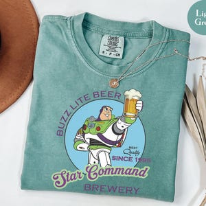 May include: Light green t-shirt featuring a cartoon image of Buzz Lightyear holding a beer mug. The shirt has the text "Buzz Lite Beer Star Command Brewery Since 1995". A silver necklace is draped across the shirt.