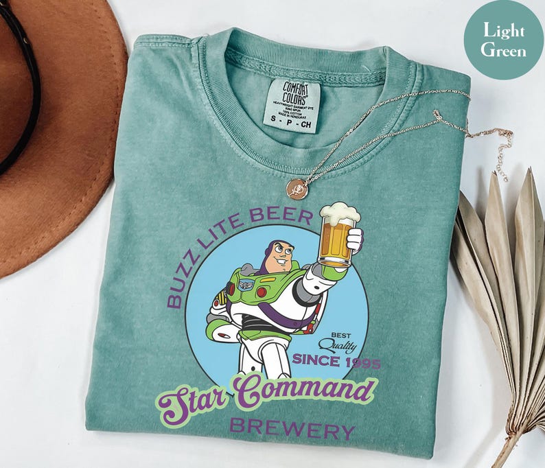 May include: Light green t-shirt featuring a cartoon image of Buzz Lightyear holding a beer mug. The shirt has the text "Buzz Lite Beer Star Command Brewery Since 1995". A silver necklace is draped across the shirt.