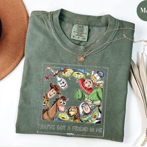 Puede incluir: Camiseta verde musgo con un gr&aacute;fico enmarcado de personajes de Toy Story, incluyendo a Woody, Buzz Lightyear y Rex. El texto "YOU'VE GOT A FRIEND IN ME" est&aacute; debajo del gr&aacute;fico. Un collar dorado est&aacute; drapeado sobre la camiseta.