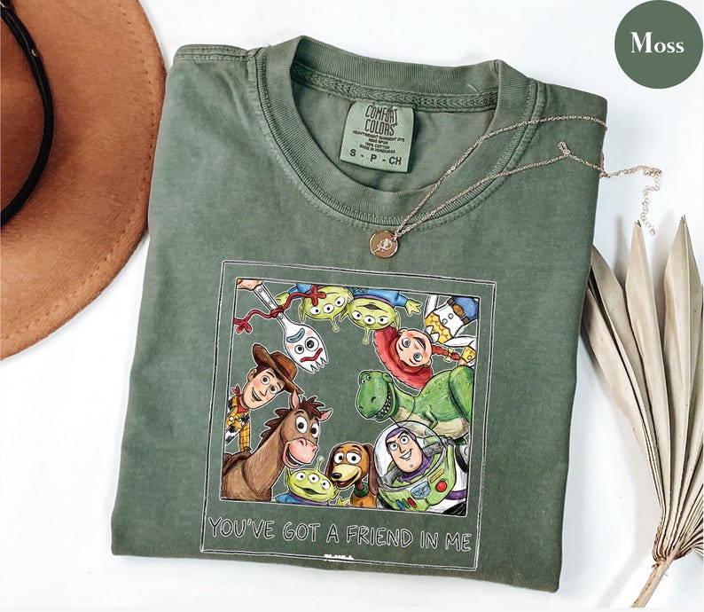 Puede incluir: Camiseta verde musgo con un gr&aacute;fico enmarcado de personajes de Toy Story, incluyendo a Woody, Buzz Lightyear y Rex. El texto "YOU'VE GOT A FRIEND IN ME" est&aacute; debajo del gr&aacute;fico. Un collar dorado est&aacute; drapeado sobre la camiseta.