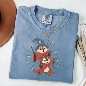 May include: A light blue t-shirt featuring a cartoon graphic of two chipmunks dressed as wizards, with a magic wand and stars. The shirt has a crew neck and short sleeves. A gold necklace is draped over the shirt.