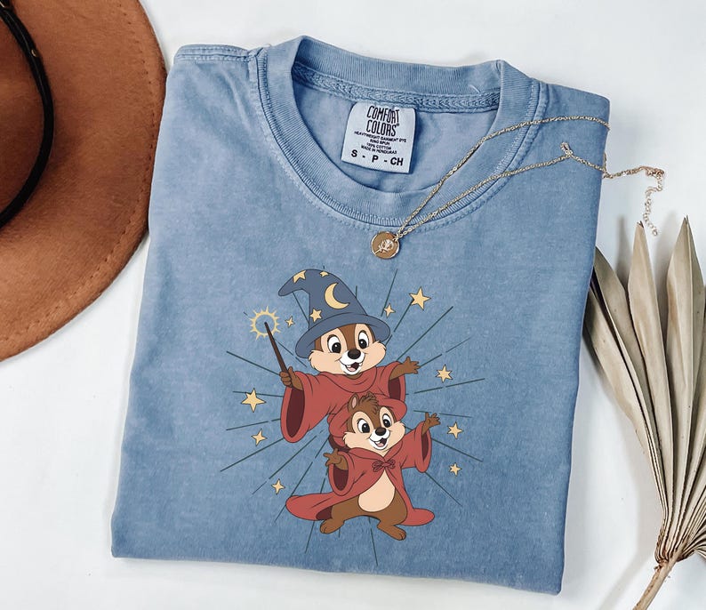 May include: A light blue t-shirt featuring a cartoon graphic of two chipmunks dressed as wizards, with a magic wand and stars. The shirt has a crew neck and short sleeves. A gold necklace is draped over the shirt.