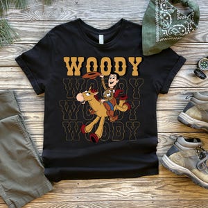 May include: Black t-shirt featuring a graphic of Woody from Toy Story riding a horse, with the word "WOODY" in a retro font. The shirt is displayed with khaki pants, hiking boots, and a green bandana on a wooden surface.