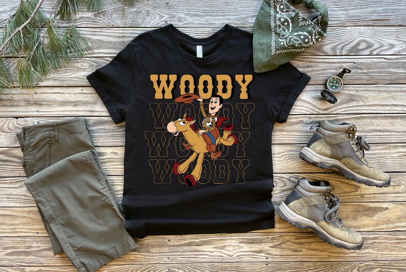 May include: Black t-shirt featuring a graphic of Woody from Toy Story riding a horse, with the word "WOODY" in a retro font. The shirt is displayed with khaki pants, hiking boots, and a green bandana on a wooden surface.