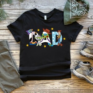 May include: Black t-shirt featuring the word "DAD" in a colourful design with Toy Story characters. The image also shows grey trousers, boots, a green bandana, and a compass, all arranged on a wooden surface.
