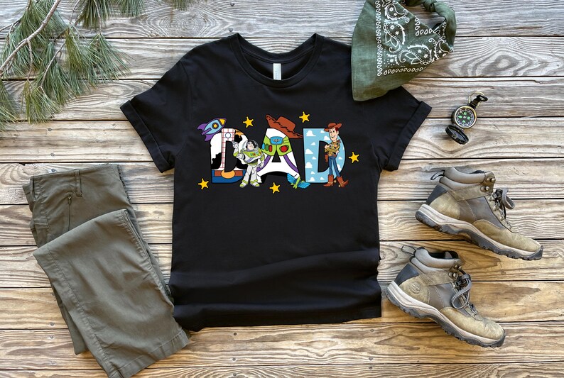 May include: Black t-shirt featuring the word "DAD" in a colourful design with Toy Story characters. The image also shows grey trousers, boots, a green bandana, and a compass, all arranged on a wooden surface.