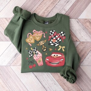 Puede incluir: Sudadera verde con un dise&ntilde;o gr&aacute;fico que presenta un coche de carreras rojo, un batido y otros elementos tem&aacute;ticos de carreras. El dise&ntilde;o incluye el texto "KA-CHOW" en galletas en forma de coraz&oacute;n y una bandera a cuadros. La sudadera tiene pu&ntilde;os.