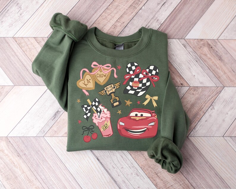 Puede incluir: Sudadera verde con un dise&ntilde;o gr&aacute;fico que presenta un coche de carreras rojo, un batido y otros elementos tem&aacute;ticos de carreras. El dise&ntilde;o incluye el texto "KA-CHOW" en galletas en forma de coraz&oacute;n y una bandera a cuadros. La sudadera tiene pu&ntilde;os.