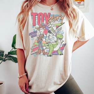 K&ouml;nnte beinhalten: Cremefarbenes T-Shirt mit farbenfroher Grafik von Charakteren aus dem Film "Toy Story". Das Design zeigt Woody, Buzz Lightyear und Rex, mit den Worten "Toy Story" und "Seit 1995."