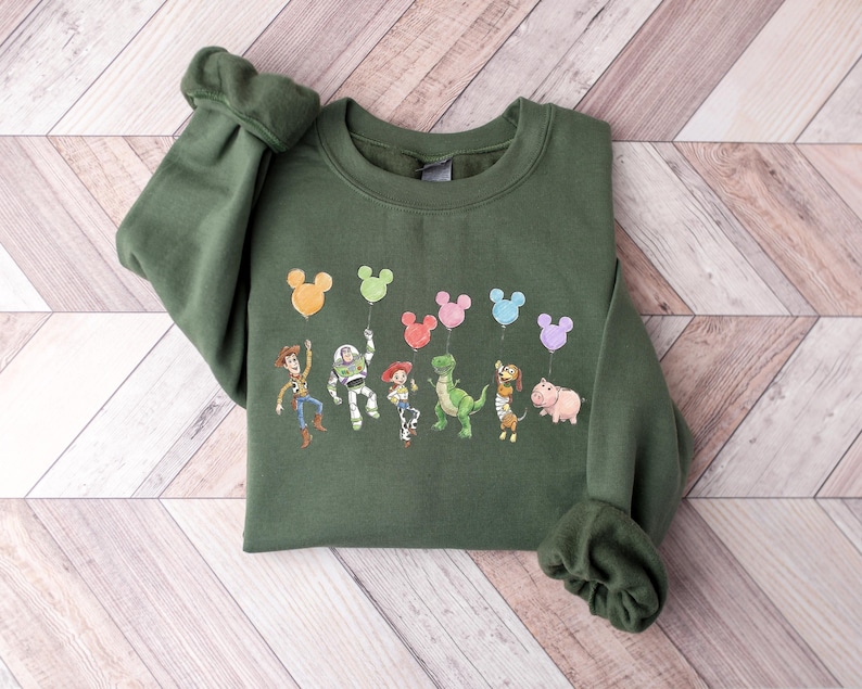 Puede incluir: Una sudadera de cuello redondo color verde oliva con un estampado gr&aacute;fico de seis personajes animados de Toy Story. Cada personaje sostiene una cuerda atada a un globo de colores con forma de Mickey Mouse. Los personajes incluyen a Woody, Buzz Lightyear, Jessie, Rex, Slinky Dog y Hamm.