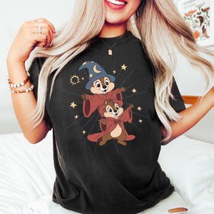 May include: Black t-shirt featuring a cartoon graphic of two chipmunks dressed as wizards, one wearing a blue hat with a crescent moon and stars. The chipmunks are wearing red robes and surrounded by gold stars and a sunburst design.