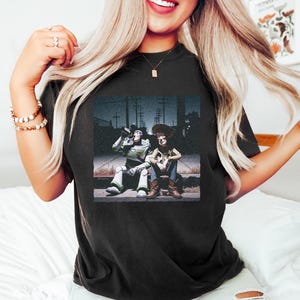 May include: A black short-sleeve t-shirt featuring a graphic print of the characters Buzz Lightyear and Woody sitting together on a curb at night. Buzz Lightyear is drinking from a bottle while Woody sits beside him in a dark outdoor setting.