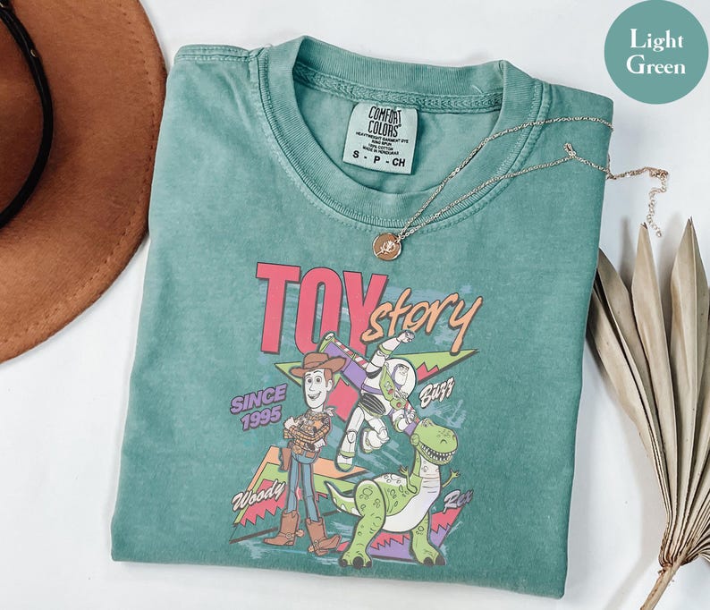 K&ouml;nnte beinhalten: Hellgr&uuml;nes T-Shirt mit einem Grafikdesign aus dem Film "Toy Story". Das Design zeigt Woody, Buzz Lightyear und Rex, mit dem Text "TOY STORY" und "Since 1995". Eine silberne Halskette mit Anh&auml;nger liegt auf dem Shirt.