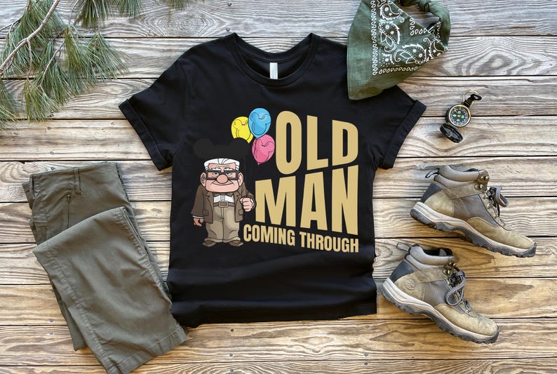 Op de afbeelding: Zwart t-shirt met de tekst "OLD MAN COMING THROUGH" en een cartoonillustratie van een oude man met ballonnen. De afbeelding bevat een kaki broek, wandelschoenen en een groene bandana.