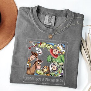 Puede incluir: Una camiseta gris Comfort Colors con un gr&aacute;fico colorido de personajes de Toy Story, incluyendo a Woody y Buzz Lightyear. El texto "YOU'VE GOT A FRIEND IN ME" est&aacute; impreso debajo. Un collar dorado est&aacute; sobre la camiseta. Talla S.