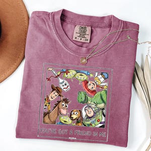 Puede incluir: Una camiseta Comfort Colors malva con un gr&aacute;fico colorido de personajes de Toy Story. El gr&aacute;fico est&aacute; enmarcado con un borde blanco e incluye el texto "You've Got a Friend in Me". Un collar de oro con un colgante est&aacute; sobre la camiseta.