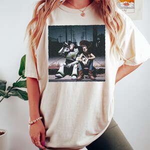 May include: A cream-colored short-sleeve t-shirt featuring a graphic print of the characters Buzz Lightyear and Woody from Toy Story sitting on a curb at night. The graphic shows Buzz Lightyear drinking from a bottle while Woody sits beside him.