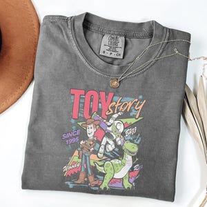 K&ouml;nnte beinhalten: Ein anthrazitfarbenes T-Shirt mit einer farbenfrohen Grafik der Toy Story-Charaktere Woody, Buzz Lightyear und Rex. Der Text "TOY STORY" ist in Rosa und Blau gehalten, darunter "SINCE 1995". Eine silberne Halskette mit Anh&auml;nger liegt auf dem Shirt.