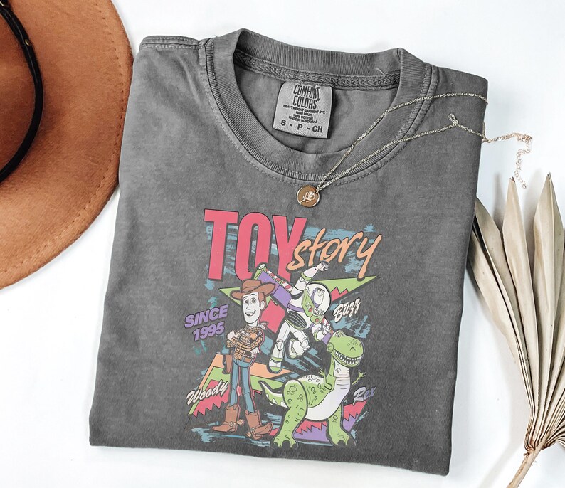 K&ouml;nnte beinhalten: Ein anthrazitfarbenes T-Shirt mit einer farbenfrohen Grafik der Toy Story-Charaktere Woody, Buzz Lightyear und Rex. Der Text "TOY STORY" ist in Rosa und Blau gehalten, darunter "SINCE 1995". Eine silberne Halskette mit Anh&auml;nger liegt auf dem Shirt.
