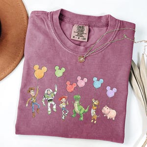 May include: A mauve short-sleeved t-shirt featuring a graphic print of six animated characters holding colourful Mickey Mouse-shaped balloons. The inner neck label reads Comfort Colors, 100% cotton, made in Honduras, size S-CH. A gold-toned necklace rests on the fabric.