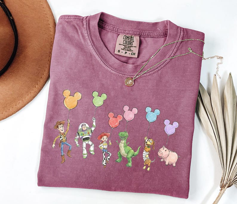 May include: A mauve short-sleeved t-shirt featuring a graphic print of six animated characters holding colourful Mickey Mouse-shaped balloons. The inner neck label reads Comfort Colors, 100% cotton, made in Honduras, size S-CH. A gold-toned necklace rests on the fabric.