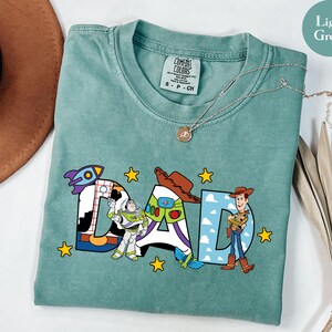 May include: Light green t-shirt featuring the word "DAD" in colourful cartoon lettering. The letters are decorated with images of characters from a popular animated film. The shirt has a crew neck and short sleeves.