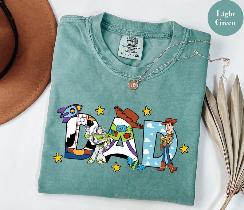 May include: Light green t-shirt featuring the word "DAD" in colourful cartoon lettering. The letters are decorated with images of characters from a popular animated film. The shirt has a crew neck and short sleeves.
