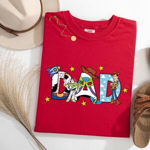 May include: A red t-shirt with the word "DAD" in colourful letters, each decorated with characters from the Toy Story movie. A rocket, cowboy hat, and Woody are featured. A gold necklace, sandals, and a hat are also in the image.