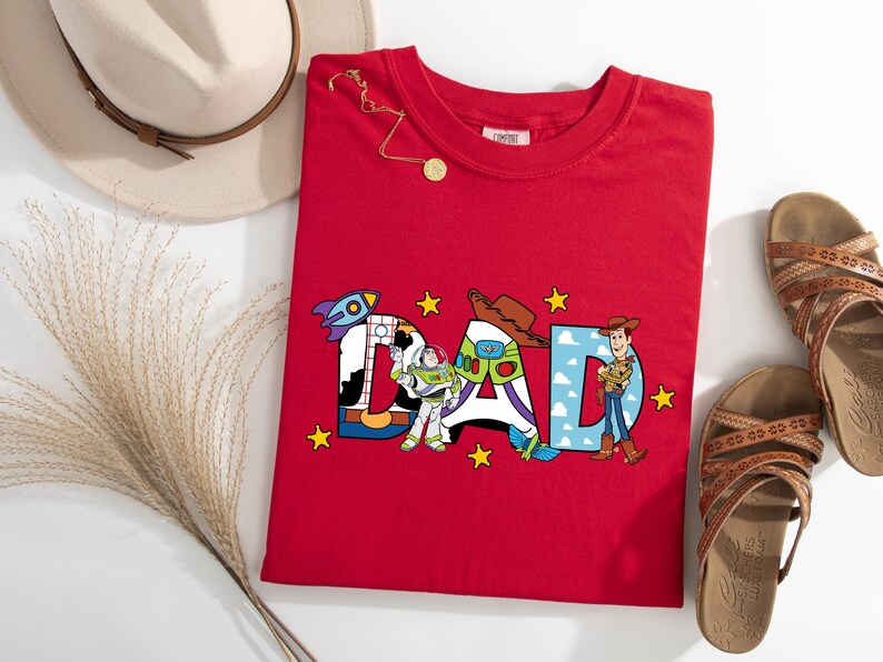 May include: A red t-shirt with the word "DAD" in colourful letters, each decorated with characters from the Toy Story movie. A rocket, cowboy hat, and Woody are featured. A gold necklace, sandals, and a hat are also in the image.