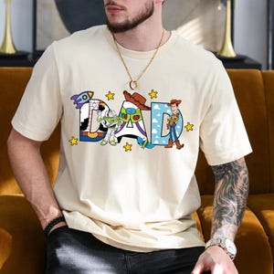 May include: A cream-coloured t-shirt featuring the word "DAD" in a colourful, cartoon font. The letters are decorated with characters from the Toy Story film, with yellow star accents. The shirt is a casual, short-sleeved design.