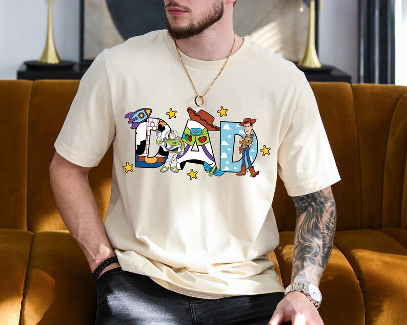 May include: A cream-coloured t-shirt featuring the word "DAD" in a colourful, cartoon font. The letters are decorated with characters from the Toy Story film, with yellow star accents. The shirt is a casual, short-sleeved design.