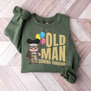 Op de afbeelding: Groen sweatshirt met de tekst "OLD MAN COMING THROUGH" in goud. Het ontwerp bevat een cartoon illustratie van een oude man die ballonnen vasthoudt. Het sweatshirt ligt op een licht houtnerf oppervlak.