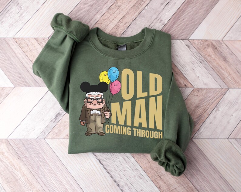 Op de afbeelding: Groen sweatshirt met de tekst "OLD MAN COMING THROUGH" in goud. Het ontwerp bevat een cartoon illustratie van een oude man die ballonnen vasthoudt. Het sweatshirt ligt op een licht houtnerf oppervlak.