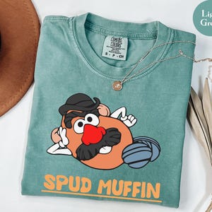 Puede incluir: Camiseta verde claro con un dise&ntilde;o de dibujos animados de Mr. Potato Head. El personaje tiene un sombrero negro, una nariz roja y un bigote negro. Las palabras "SPUD MUFFIN" est&aacute;n impresas en naranja.