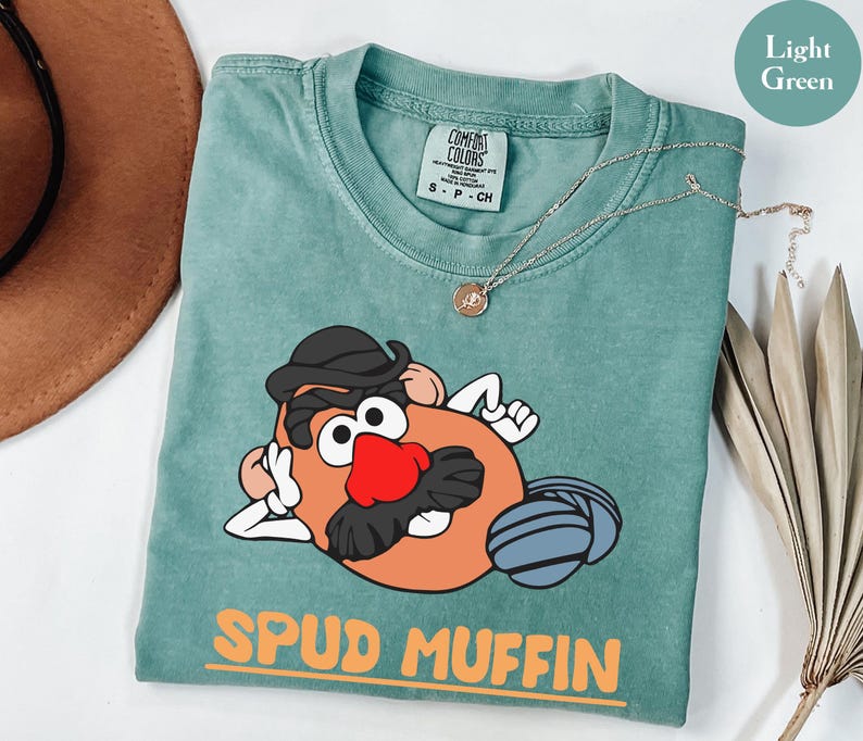 Puede incluir: Camiseta verde claro con un dise&ntilde;o de dibujos animados de Mr. Potato Head. El personaje tiene un sombrero negro, una nariz roja y un bigote negro. Las palabras "SPUD MUFFIN" est&aacute;n impresas en naranja.