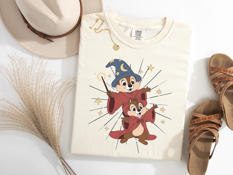 May include: Cream-colored t-shirt featuring a cartoon wizard design with two chipmunks wearing wizard robes and hats. The design includes stars and a magic wand. The shirt is styled with a hat, sandals, and jewelry.