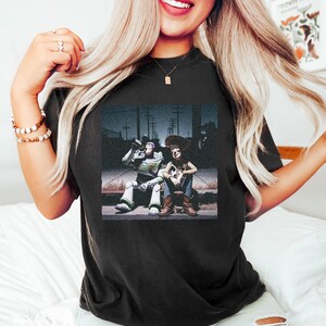 May include: A black t-shirt featuring a graphic print of the characters Buzz Lightyear and Woody sitting together on a curb at night. The design shows Buzz Lightyear in his space suit and Woody in his cowboy outfit, set against a dark street background.
