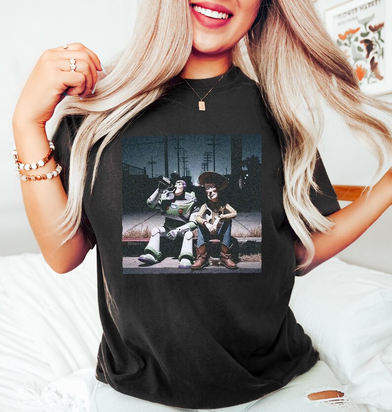 May include: A black t-shirt featuring a graphic print of the characters Buzz Lightyear and Woody sitting together on a curb at night. The design shows Buzz Lightyear in his space suit and Woody in his cowboy outfit, set against a dark street background.
