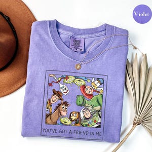 Puede incluir: Una camiseta violeta Comfort Colors con un gr&aacute;fico enmarcado de personajes de Toy Story, incluyendo a Woody y Buzz Lightyear. El texto "YOU'VE GOT A FRIEND IN ME" rodea el gr&aacute;fico. Un collar dorado est&aacute; sobre la camiseta. La camiseta es de color violeta.