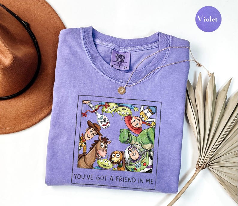 Puede incluir: Una camiseta violeta Comfort Colors con un gr&aacute;fico enmarcado de personajes de Toy Story, incluyendo a Woody y Buzz Lightyear. El texto "YOU'VE GOT A FRIEND IN ME" rodea el gr&aacute;fico. Un collar dorado est&aacute; sobre la camiseta. La camiseta es de color violeta.