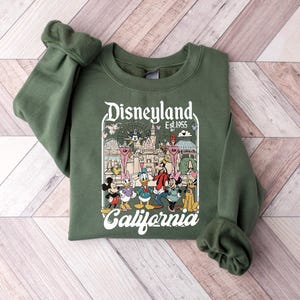 May include: An olive green crewneck sweatshirt featuring a graphic print of Disneyland California Est. 1955. The design includes illustrations of Mickey Mouse, Minnie Mouse, Donald Duck, Daisy Duck, Goofy, and Pluto in front of the castle.