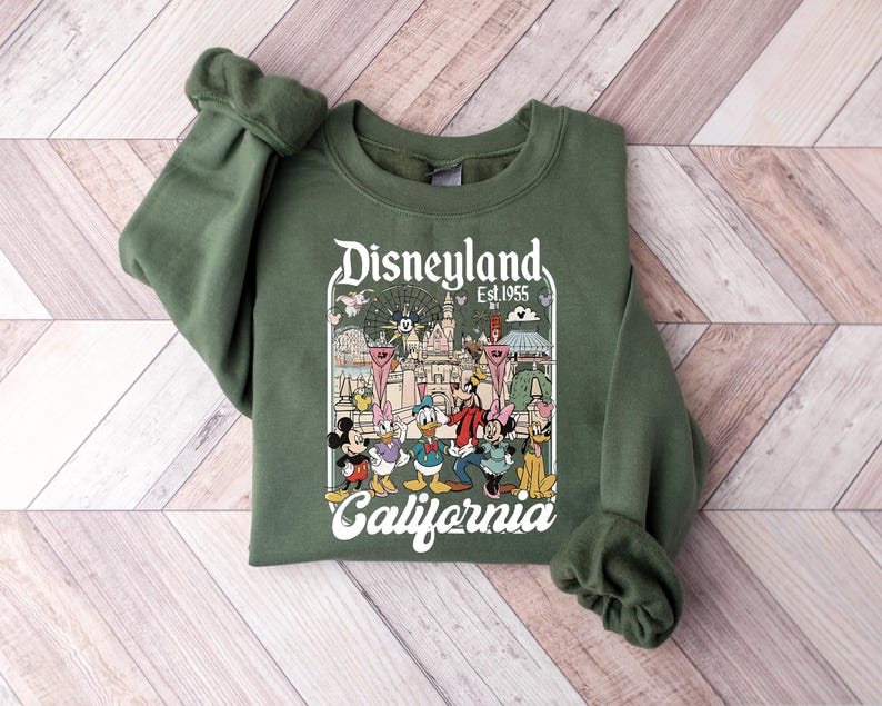 May include: An olive green crewneck sweatshirt featuring a graphic print of Disneyland California Est. 1955. The design includes illustrations of Mickey Mouse, Minnie Mouse, Donald Duck, Daisy Duck, Goofy, and Pluto in front of the castle.