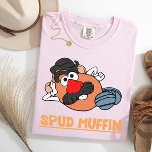 Puede incluir: Camiseta rosa claro con un personaje de dibujos animados de patata con sombrero y bigote, y el texto "SPUD MUFFIN" en naranja. La camiseta se presenta con sandalias, un sombrero y un collar.