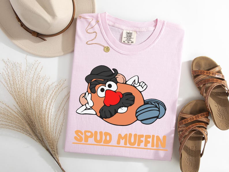 Puede incluir: Camiseta rosa claro con un personaje de dibujos animados de patata con sombrero y bigote, y el texto "SPUD MUFFIN" en naranja. La camiseta se presenta con sandalias, un sombrero y un collar.