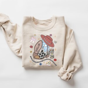 K&ouml;nnte beinhalten: Ein beigefarbenes Sweatshirt mit Rundhalsausschnitt und einem Grafikdruck, der einen Cowboystiefel mit Kuhfleckenmuster, einen roten Cowboyhut, eine Schleife und W&uuml;rfel mit den Buchstaben T, S und S zeigt. Das Wort TAYLOR steht auf der Sohle des Stiefels und die Zahl 13 ist auf dem Stiefelschaft zu sehen.