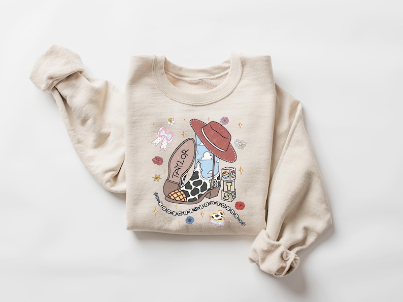 K&ouml;nnte beinhalten: Ein beigefarbenes Sweatshirt mit Rundhalsausschnitt und einem Grafikdruck, der einen Cowboystiefel mit Kuhfleckenmuster, einen roten Cowboyhut, eine Schleife und W&uuml;rfel mit den Buchstaben T, S und S zeigt. Das Wort TAYLOR steht auf der Sohle des Stiefels und die Zahl 13 ist auf dem Stiefelschaft zu sehen.