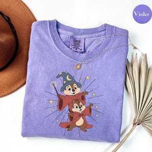 May include: A violet Comfort Colors crew neck t-shirt featuring a graphic of two cartoon chipmunks dressed as wizards. One chipmunk wears a blue wizard hat and holds a wand. A gold necklace and a brown hat are also visible.