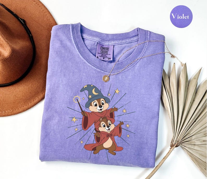 May include: A violet Comfort Colors crew neck t-shirt featuring a graphic of two cartoon chipmunks dressed as wizards. One chipmunk wears a blue wizard hat and holds a wand. A gold necklace and a brown hat are also visible.