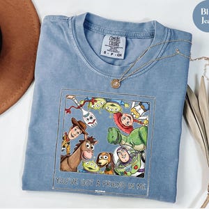 Puede incluir: Camiseta azul vaquero Comfort Colors con un gr&aacute;fico colorido de personajes de una popular pel&iacute;cula animada. El gr&aacute;fico est&aacute; enmarcado con el texto "YOU'VE GOT A FRIEND IN ME". Un collar dorado est&aacute; sobre la camisa.