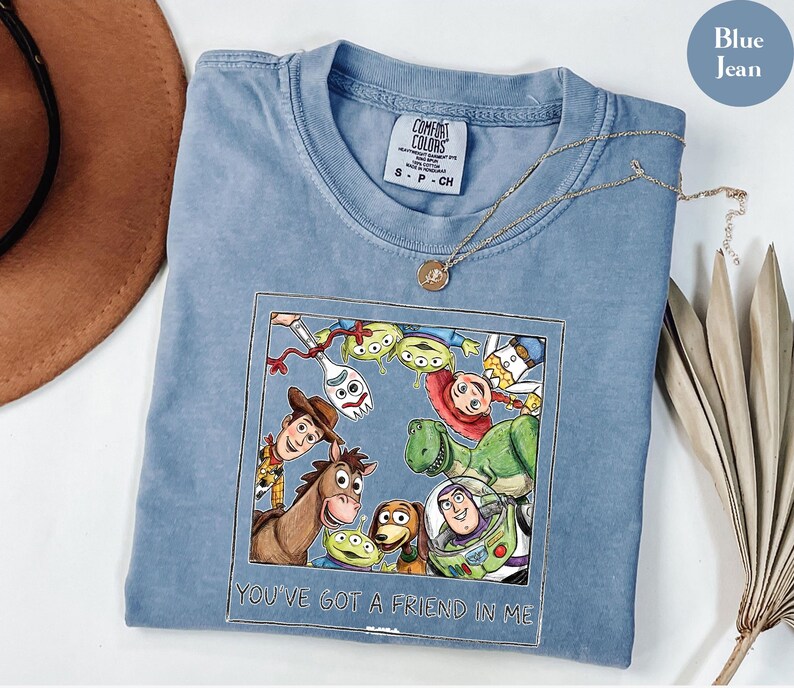 Puede incluir: Camiseta azul vaquero Comfort Colors con un gr&aacute;fico colorido de personajes de una popular pel&iacute;cula animada. El gr&aacute;fico est&aacute; enmarcado con el texto "YOU'VE GOT A FRIEND IN ME". Un collar dorado est&aacute; sobre la camisa.